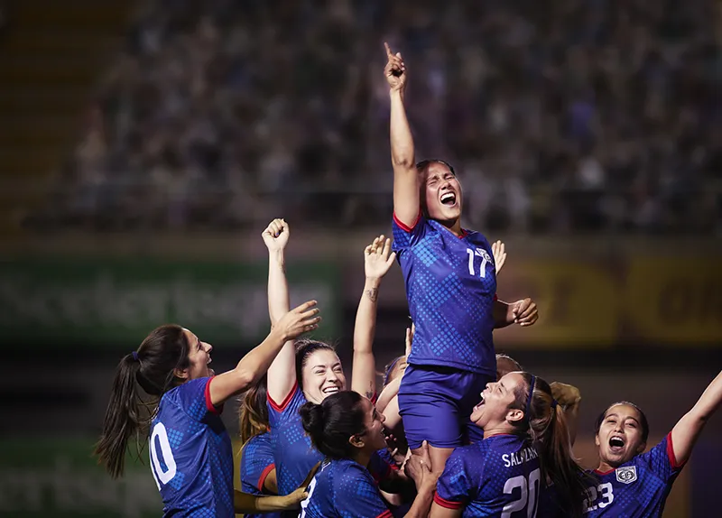 Women's soccer team celebrating a victory, lifting a teammate wearing jersey number 17 who is raising her fist triumphantly under stadium lights.