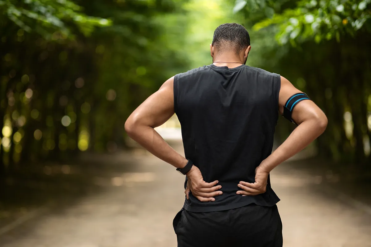 Man standing on a forest trail holding his lower back, indicating back pain or discomfort, symbolizing chronic pain.